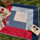 Colorful quilt on grass with picnic items including a basket, apples, sandwiches, and a book.