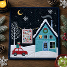 Quilted Christmas scene with a house, car, and tree on a wooden surface.