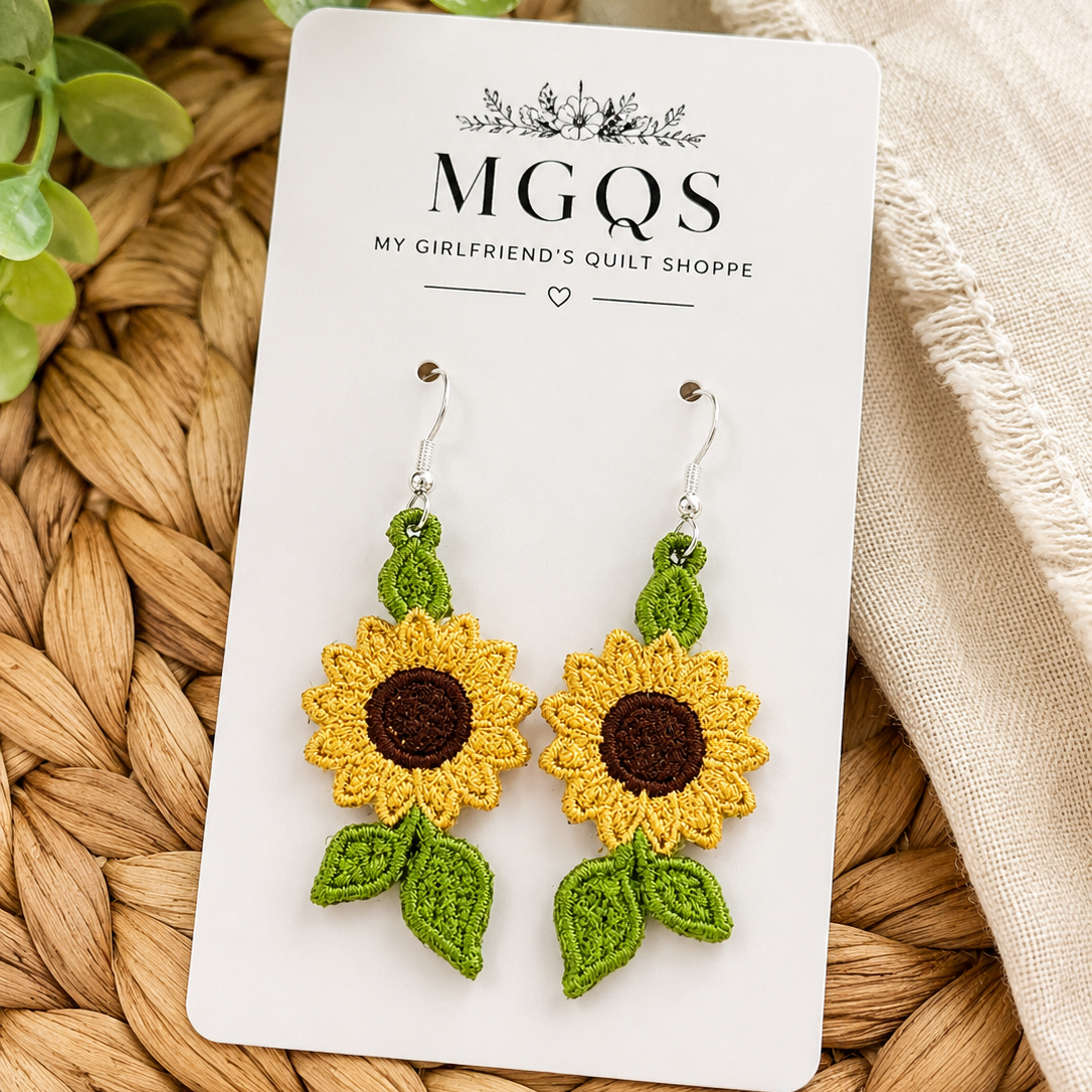 A pair of handmade crochet sunflower dangle earrings on a white display card from MGQS, styled on a woven straw mat with green leaves and a beige fabric.