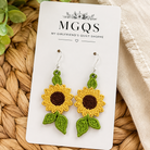 A pair of handmade crochet sunflower dangle earrings on a white display card from MGQS, styled on a woven straw mat with green leaves and a beige fabric.