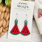 Watermelon slice embroidered patch earrings on a white display card by MGQS, styled on a woven mat with a printed fabric napkin and green leaves.