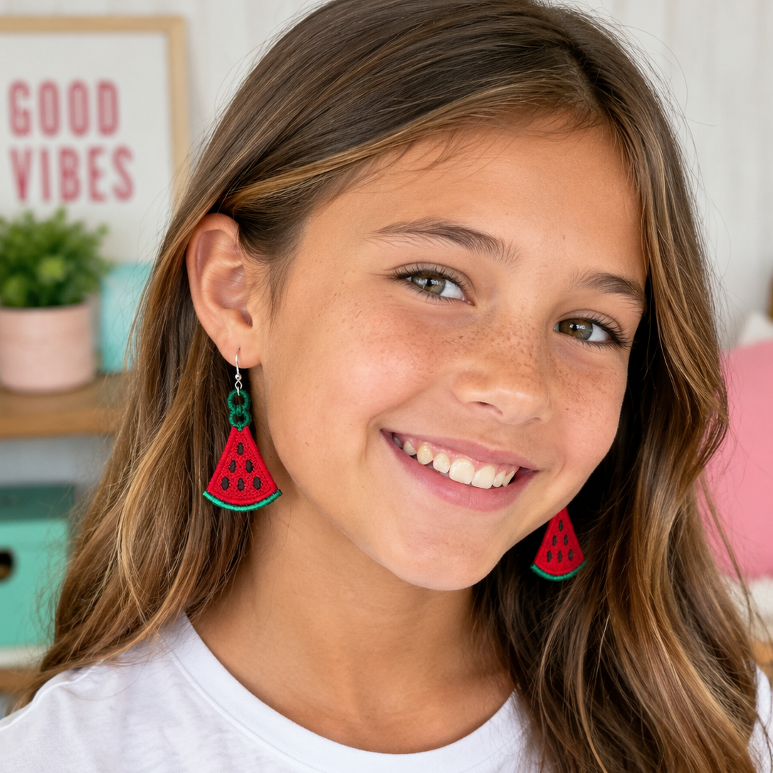 A smiling young girl with long brown hair wearing watermelon-shaped dangling earrings, photographed in a bright indoor setting.